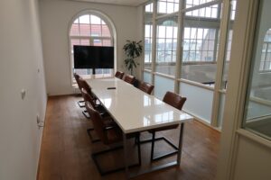 Vergaderruimte aan het Gedempt Hamerkanaal met lange witte tafel, bruine stoelen, televisie en grote ramen met stadsuitzicht.